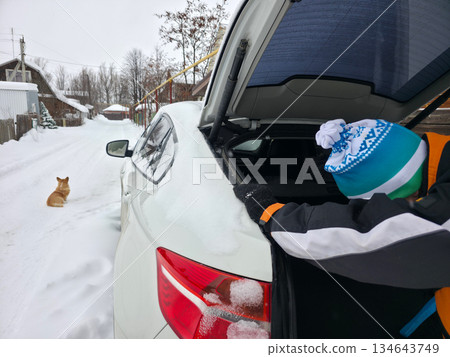 Man cleaning snow from car trunk in winter village street, with a dog sitting nearby. Cold season and travel preparation. Man cleaning snow from car trunk in winter village street, with a dog sitting nearby. Cold season and travel preparation. 134643749