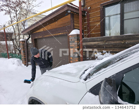 Man brushing snow off white car in winter weather, concept of vehicle maintenance and cold season challenges, cleaning, automobile care. 134643752