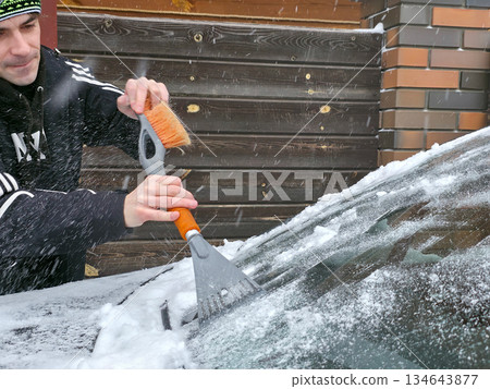 Man removing snow from car windshield with an ice scraper and brush. Winter car maintenance and preparation for cold weather. Man removing snow from car windshield with an ice scraper and brush. Winter car maintenance and preparation for cold weather. 134643877