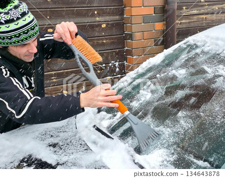 Man scraping snow from car windshield with an ice scraper and brush. Winter concept for cold weather, vehicle maintenance, and daily commute preparation. 134643878