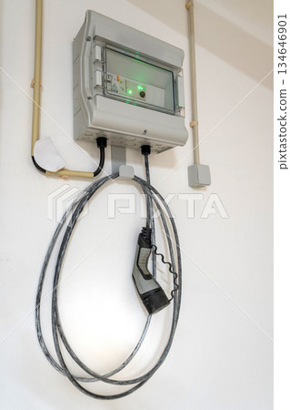 A new electricity meter is mounted on wall, displaying energy usage with a green light. Cables run down, connecting to the meter and hanging nearby. charger for electric car 134646901