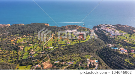 Aerial view of Falesia cliffs in Albufeira with scattered villas, green hills and winding roads leading to Atlantic Ocean boat sailing in Algarve, Portugal. Concept of rural coastal serenity Aerial view of Falesia cliffs in Albufeira with scattered villas, green hills and winding roads leading to Atlantic Ocean boat sailing in Algarve, Portugal. Concept of rural coastal serenity 134646902