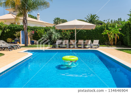 Families enjoy a sunny afternoon by the pool, featuring lounge chairs and a large umbrella in a vibrant garden filled with greenery Families enjoy a sunny afternoon by the pool, featuring lounge chairs and a large umbrella in a vibrant garden filled with greenery 134646904