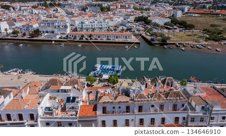 Aerial view of Tavira town with traditional tiled rooftops and boats along the Gilao River under bright sunlight in Algarve, Portugal. Concept of historic riverside tourism 134646910
