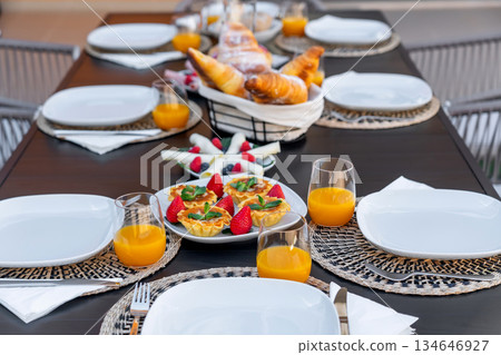 A beautifully arranged outdoor breakfast table features fresh croissants, fruit tarts, and orange juice. It's a delightful setting for a cozy morning gathering A beautifully arranged outdoor breakfast table features fresh croissants, fruit tarts, and orange juice. It's a delightful setting for a cozy morning gathering 134646927