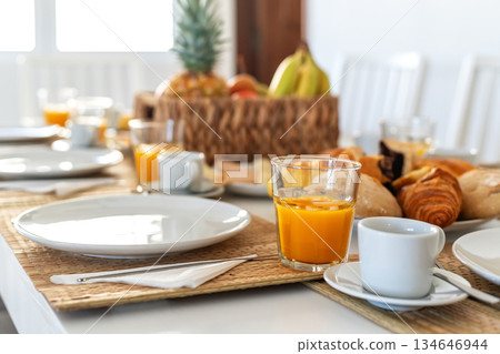 A table is set for breakfast featuring orange juice, fresh fruit, and a variety of pastries. Natural light fills the room, creating a warm, inviting atmosphere 134646944
