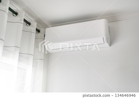 A wall-mounted air conditioner is installed in a bright room with white walls and sheer curtains, creating a cool atmosphere on a warm day 134646946