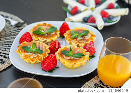Tarts filled with creamy custard are beautifully arranged with fresh strawberries. A colorful fruit dish and drinks complete the inviting breakfast spread 134646949