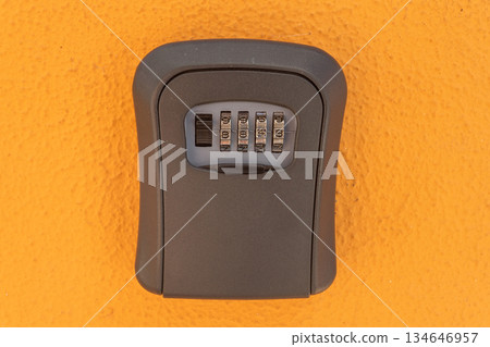 A gray lockbox with a combination dial is attached to a vibrant orange wall, providing a secure place to store keys for easy access 134646957