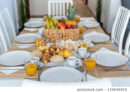 A beautifully arranged breakfast table features fresh fruits, various pastries, and beverages in a bright, modern dining space 134646958