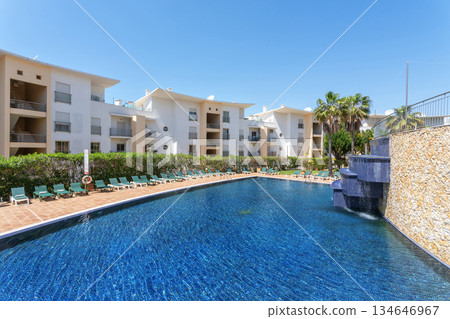 Visitors enjoy a serene day by the sparkling pool at a resort. The bright blue water contrasts with the surrounding greenery and modern architecture on a sunny afternoon Visitors enjoy a serene day by the sparkling pool at a resort. The bright blue water contrasts with the surrounding greenery and modern architecture on a sunny afternoon 134646967