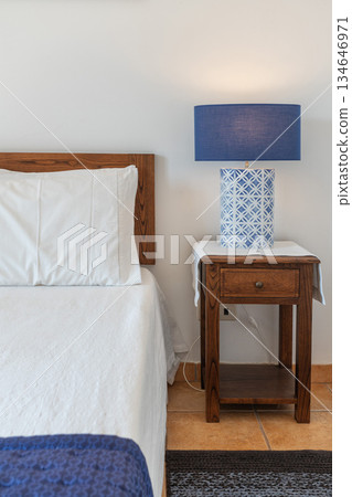 Soft white bedding enhances the inviting atmosphere of this cozy bedroom featuring a wooden nightstand and a stylish blue lamp on the side Soft white bedding enhances the inviting atmosphere of this cozy bedroom featuring a wooden nightstand and a stylish blue lamp on the side 134646971