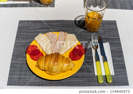 A colorful plate features a flaky croissant, slices of cheese, turkey, and pepperoni. A glass of orange juice sits on the table, perfect for breakfast A colorful plate features a flaky croissant, slices of cheese, turkey, and pepperoni. A glass of orange juice sits on the table, perfect for breakfast 134646973