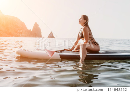 Woman Paddleboard Ocean Coastline - A woman in a swimsuit sits on a paddleboard in the ocean, enjoying the view of the coastline. 134648232