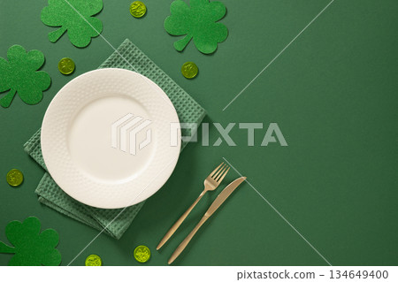 Festive St Patricks day table setting with white plate, gold cutlery and green shamrock decorations on green background. Elegant holiday mockup with copy space. View from above. Festive St Patricks day table setting with white plate, gold cutlery and green shamrock decorations on green background. Elegant holiday mockup with copy space. View from above. 134649400
