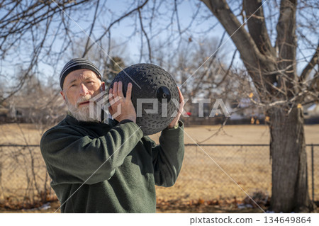 senior man is exercising with a heavy, 50 lb, slam ball in his backyard, sunny winter day senior man is exercising with a heavy, 50 lb, slam ball in his backyard, sunny winter day 134649864