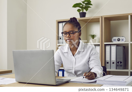 Attentive young african-american girl with glasses, dressed in a white shirt is sitting at her desk. Attentive young african-american girl with glasses, dressed in a white shirt is sitting at her desk. 134649871