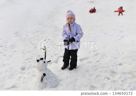 A girl in a hat and gloves makes a snowman in the snow A girl in a hat and gloves makes a snowman in the snow 134650172