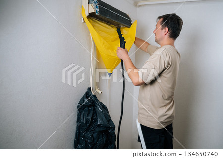 Professional technician male cleaning air conditioning unit using steam cleaner with protective sheet catching debris. Concept for maintenance of hygiene and pure air to prevent allergies Professional technician male cleaning air conditioning unit using steam cleaner with protective sheet catching debris. Concept for maintenance of hygiene and pure air to prevent allergies 134650740