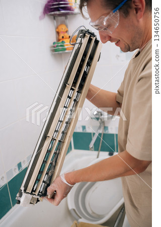 Smiling professional HVAC technician man cleaning dirty air conditioner parts with detergent, ensuring proper hygiene and improved air quality. Concept of air condition maintenance service. Smiling professional HVAC technician man cleaning dirty air conditioner parts with detergent, ensuring proper hygiene and improved air quality. Concept of air condition maintenance service. 134650756