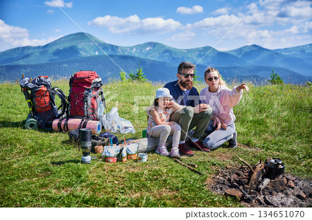Family of three enjoys camping trip in mountains, with backpacks and camping gear around. Mother points at something in distance while father and daughter, taking in scenic view of lush green hills. Family of three enjoys camping trip in mountains, with backpacks and camping gear around. Mother points at something in distance while father and daughter, taking in scenic view of lush green hills. 134651070