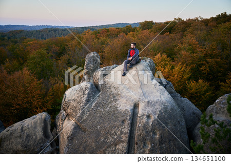 Aerial view of climber sitting atop rock formation at Dovbush Rocks in Carpathian mountains, Ukraine. Sun sets, casting warm glow over autumn forest and distant hills, creating breathtaking view. Aerial view of climber sitting atop rock formation at Dovbush Rocks in Carpathian mountains, Ukraine. Sun sets, casting warm glow over autumn forest and distant hills, creating breathtaking view. 134651100