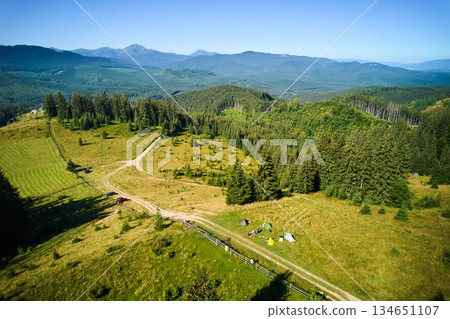 Aerial view campsite in mountains, with several colorful tents on grassy clearing. Campers relax under clear blue sky, surrounded by tall evergreens and panoramic views of distant peaks. 134651107