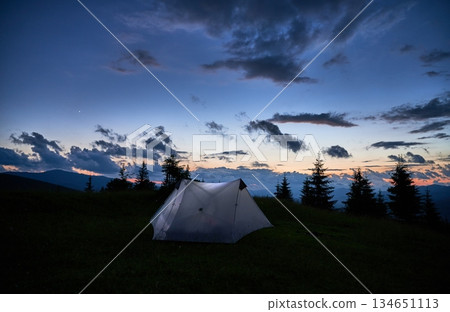 Touristic tent glows warmly on grassy hillside at dusk, surrounded by dark silhouettes of mountains and pine trees. Sky is deep blue with scattered clouds, as last light of sunset fades into night. Touristic tent glows warmly on grassy hillside at dusk, surrounded by dark silhouettes of mountains and pine trees. Sky is deep blue with scattered clouds, as last light of sunset fades into night. 134651113