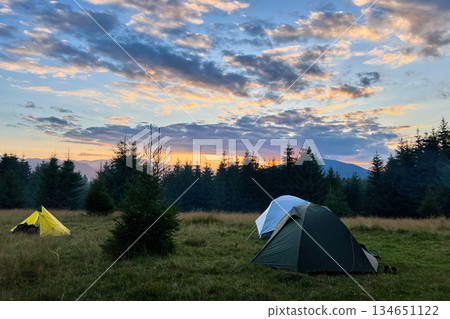 Few tourist tents in grassy field in evening, surrounded by trees and hills under stunning sunset sky. Colorful clouds enhance beauty of scene, with hues of orange and purple illuminating horizon. Few tourist tents in grassy field in evening, surrounded by trees and hills under stunning sunset sky. Colorful clouds enhance beauty of scene, with hues of orange and purple illuminating horizon. 134651122
