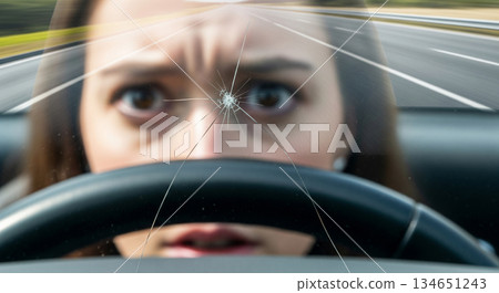 A driver looks startled as a small crack spreads quickly across the windshield. The scene captures the moment on a highway, highlighting the sudden change in visibility. 134651243