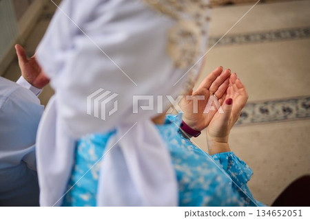 Closeup hands raised in prayer young girl wearing blue hijab kneeling on patterned carpet inside mosque delicate bracelet warm light serene focus spiritual devotion 134652051