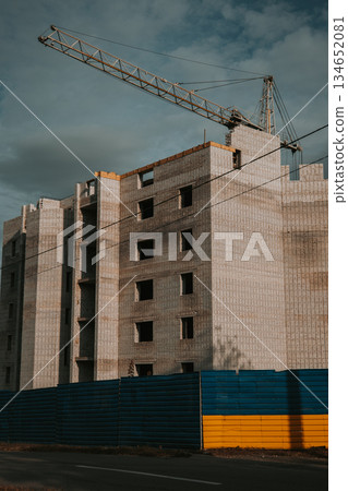 Construction of a new apartment building with a blue and yellow fence in front. The unfinished building stands behind a fence painted in the colors of the Ukrainian flag. Real estate market of Ukraine 134652081