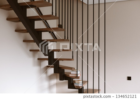 Modern floating staircase with wooden steps and black metal railings. A stack of books rests on the steps of contemporary staircase, minimalist design Modern floating staircase with wooden steps and black metal railings. A stack of books rests on the steps of contemporary staircase, minimalist design 134652121