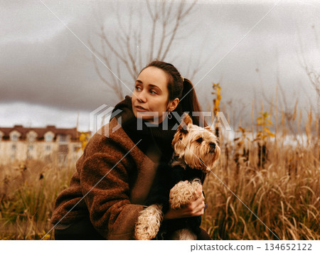 Woman embracing her small dog outdoors on an autumn day. A stylish young white woman holds her doggy, lapdog close while enjoying a walk in a park. Woman embracing her small dog outdoors on an autumn day. A stylish young white woman holds her doggy, lapdog close while enjoying a walk in a park. 134652122