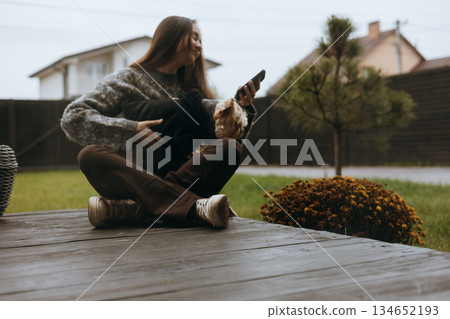 Woman and her dog enjoyving a relaxing day outdoors in the backyard patio. A young woman sits cross-legged on a wooden deck of home terrace with her small dog, enjoying the fresh air Cottage lifestyle Woman and her dog enjoyving a relaxing day outdoors in the backyard patio. A young woman sits cross-legged on a wooden deck of home terrace with her small dog, enjoying the fresh air Cottage lifestyle 134652193