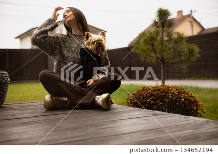 Woman and her dog enjoyving a relaxing day outdoors in the backyard patio. A young woman sits cross-legged on a wooden deck of home terrace with her small dog, enjoying the fresh air Cottage lifestyle Woman and her dog enjoyving a relaxing day outdoors in the backyard patio. A young woman sits cross-legged on a wooden deck of home terrace with her small dog, enjoying the fresh air Cottage lifestyle 134652194