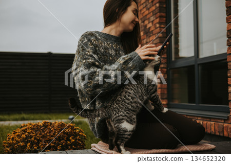 Cozy autumn scene with a woman sitting on patio terrace near a brick house, playing with her cat while holding a smartphone. Relaxed lifestyle moment. 134652320
