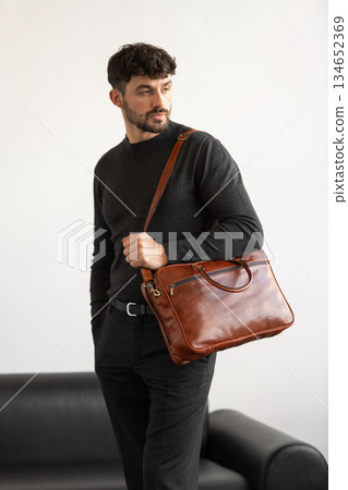 Professional male model in charcoal crewneck knitwear holding a light brown leather briefcase in a minimalist studio setting 134652369