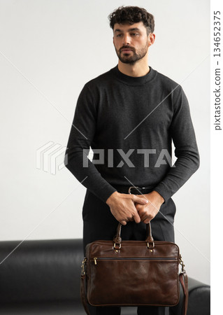 Professional male model in charcoal crewneck knitwear holding a dark brown leather briefcase in a minimalist studio setting 134652375