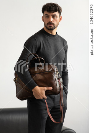 Professional male model in charcoal crewneck knitwear holding a dark brown leather briefcase in a minimalist studio setting 134652376