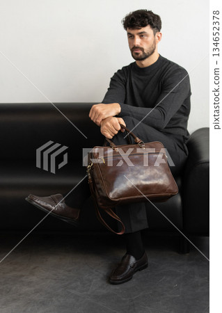A professional male model in charcoal knitwear and tailored trousers sits on a black leather sofa holding a premium brown leather briefcase. 134652378