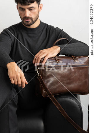 A professional male model in charcoal knitwear and tailored trousers sits on a black leather sofa holding a premium brown leather briefcase. A professional male model in charcoal knitwear and tailored trousers sits on a black leather sofa holding a premium brown leather briefcase. 134652379