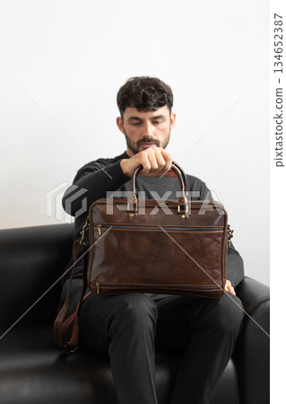 A professional male model in charcoal knitwear and tailored trousers sits on a black leather sofa holding a premium brown leather briefcase. A professional male model in charcoal knitwear and tailored trousers sits on a black leather sofa holding a premium brown leather briefcase. 134652387