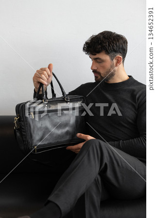 A professional male model in charcoal knitwear and tailored trousers sits on a black leather sofa holding a premium black leather briefcase. 134652391