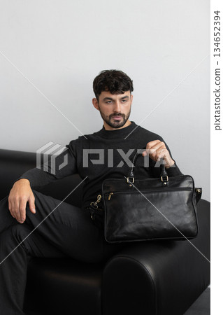 A professional male model in charcoal knitwear and tailored trousers sits on a black leather sofa holding a premium black leather briefcase. A professional male model in charcoal knitwear and tailored trousers sits on a black leather sofa holding a premium black leather briefcase. 134652394