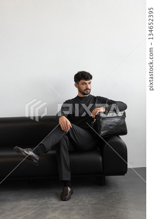 A professional male model in charcoal knitwear and tailored trousers sits on a black leather sofa holding a premium black leather briefcase. A professional male model in charcoal knitwear and tailored trousers sits on a black leather sofa holding a premium black leather briefcase. 134652395