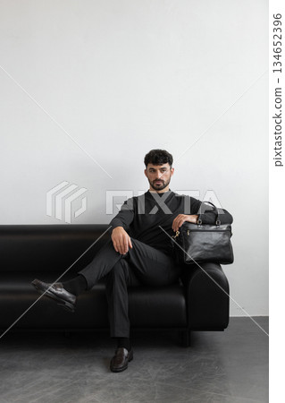 A professional male model in charcoal knitwear and tailored trousers sits on a black leather sofa holding a premium black leather briefcase. A professional male model in charcoal knitwear and tailored trousers sits on a black leather sofa holding a premium black leather briefcase. 134652396