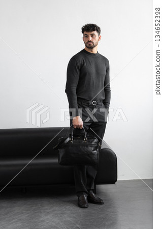A professional man in a charcoal crewneck sweater and tailored trousers holds a black leather briefcase standing in a minimalist studio. A professional man in a charcoal crewneck sweater and tailored trousers holds a black leather briefcase standing in a minimalist studio. 134652398