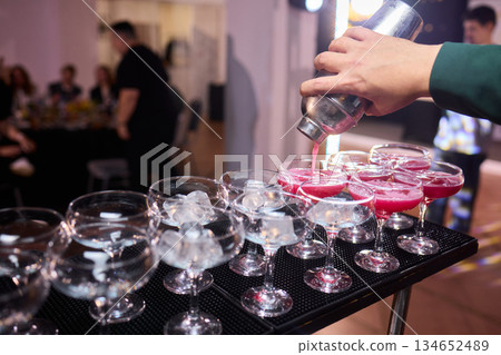 Mood setting with lively drink preparation at upscale bar, Elegant scene of bartender pouring colorful cocktail amidst festive warm lighting and guests in background 134652489