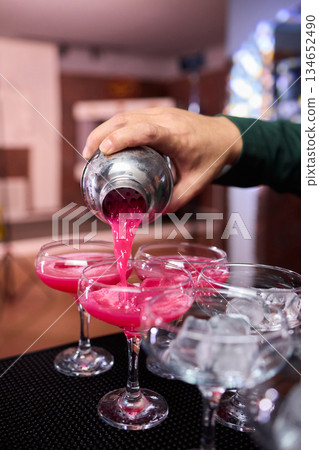 Mood setting with lively drink preparation at upscale bar, Elegant scene of bartender pouring colorful cocktail amidst festive warm lighting and guests in background 134652490